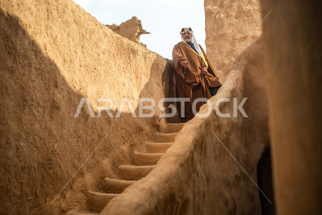 رجل مسن عربي سعودي خليجي يرتدي الزي السعودي الشعبي التقليدي, الزي التراثي للمناسبات الوطنيه, التجول في إحدى المباني الطينيه الشعبيه, بيوت تراثيه قديمه, شعبيات, أحياء أثريه قديمه, جوله بين الأحياء الشعبيه