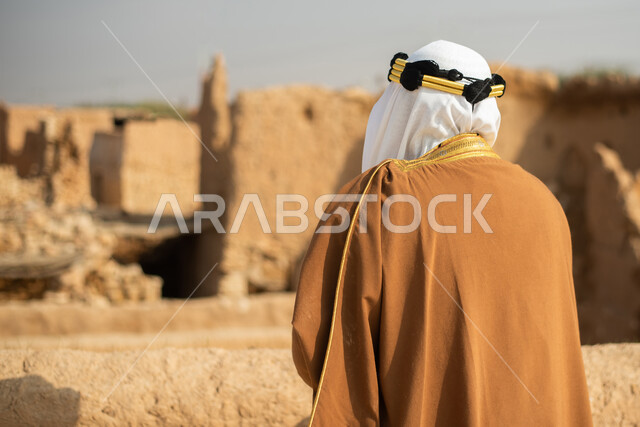 تصوير من الخلف لرجل مسن عربي سعودي خليجي يرتدي الزي السعودي الشعبي التقليدي, الزي التراثي للمناسبات الوطنيه, الوقوف في إحدى المباني الطينيه الشعبيه, رؤيه مستقبليه، المستقبل السعودي، النظر من بعيد, بيوت تراثيه, يوم التأسيس السعودي, مباني أثريه