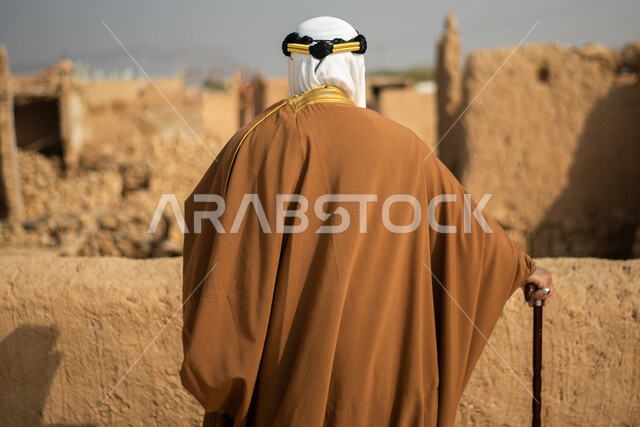 تصوير من الخلف لرجل مسن عربي سعودي خليجي يرتدي الزي السعودي الشعبي التقليدي, الزي التراثي للمناسبات الوطنيه, الوقوف في إحدى المباني الطينيه الشعبيه, رؤيه مستقبليه، المستقبل السعودي، النظر من بعيد, بيوت تراثيه, يوم التأسيس السعودي, مباني أثريه