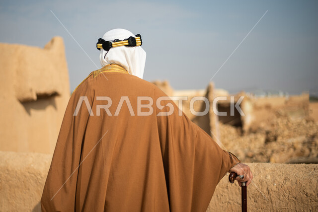 تصوير من الخلف لرجل مسن عربي سعودي خليجي يرتدي الزي السعودي الشعبي التقليدي, الزي التراثي للمناسبات الوطنيه, الوقوف في إحدى المباني الطينيه الشعبيه, رؤيه مستقبليه، المستقبل السعودي، النظر من بعيد, بيوت تراثيه, يوم التأسيس السعودي, مباني أثريه