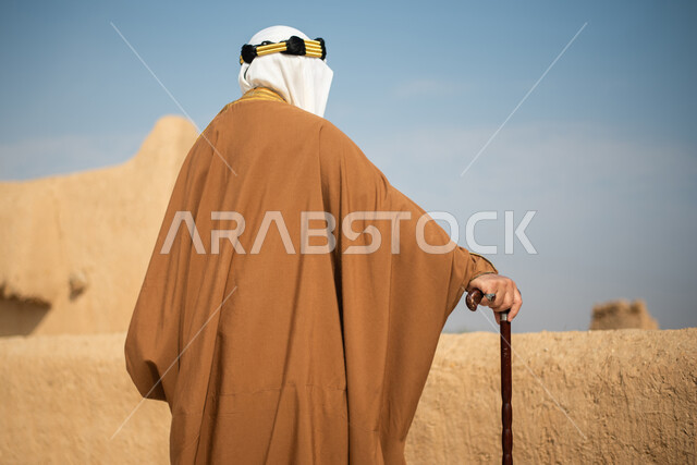 تصوير من الخلف لرجل مسن عربي سعودي خليجي يرتدي الزي السعودي الشعبي التقليدي, الزي التراثي للمناسبات الوطنيه, الوقوف في إحدى المباني الطينيه الشعبيه, رؤيه مستقبليه، المستقبل السعودي، النظر من بعيد, بيوت تراثيه, يوم التأسيس السعودي, مباني أثريه