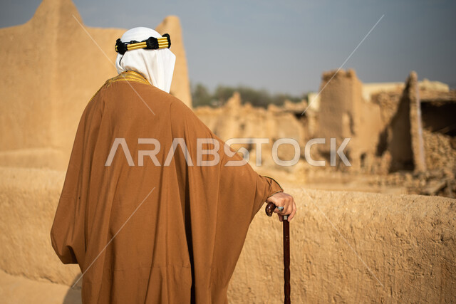 تصوير من الخلف لرجل مسن عربي سعودي خليجي يرتدي الزي السعودي الشعبي التقليدي, الزي التراثي للمناسبات الوطنيه, الوقوف في إحدى المباني الطينيه الشعبيه, رؤيه مستقبليه، المستقبل السعودي، النظر من بعيد, بيوت تراثيه, يوم التأسيس السعودي, مباني أثريه