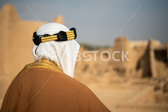 تصوير من الخلف لرجل مسن عربي سعودي خليجي يرتدي الزي السعودي الشعبي التقليدي, الزي التراثي للمناسبات الوطنيه, الوقوف في إحدى المباني الطينيه الشعبيه, رؤيه مستقبليه، المستقبل السعودي، النظر من بعيد, بيوت تراثيه, يوم التأسيس السعودي, مباني أثريه