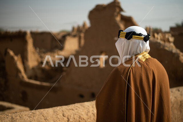 تصوير من الخلف لرجل مسن عربي سعودي خليجي يرتدي الزي السعودي الشعبي التقليدي, الزي التراثي للمناسبات الوطنيه, الوقوف في إحدى المباني الطينيه الشعبيه, رؤيه مستقبليه، المستقبل السعودي، النظر من بعيد, بيوت تراثيه, يوم التأسيس السعودي, مباني أثريه