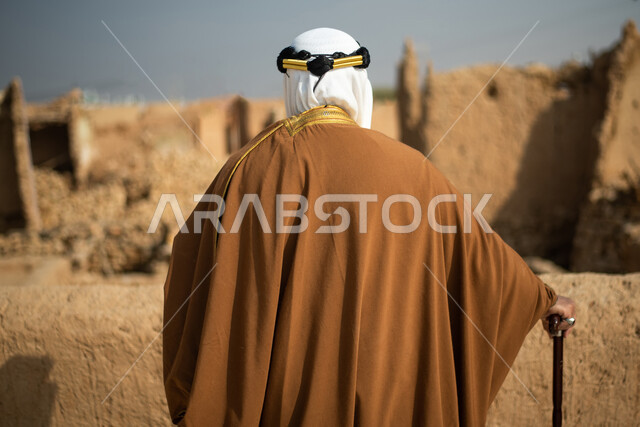 تصوير من الخلف لرجل مسن عربي سعودي خليجي يرتدي الزي السعودي الشعبي التقليدي, الزي التراثي للمناسبات الوطنيه, الوقوف في إحدى المباني الطينيه الشعبيه, رؤيه مستقبليه، المستقبل السعودي، النظر من بعيد, بيوت تراثيه, يوم التأسيس السعودي, مباني أثريه