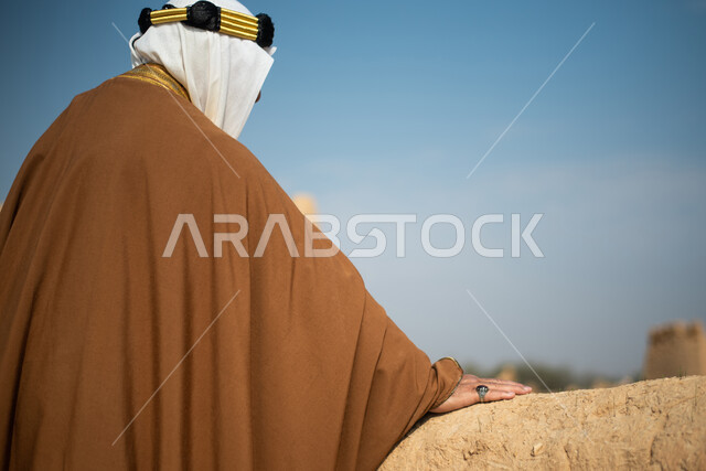 تصوير من الخلف لرجل مسن عربي سعودي خليجي يرتدي الزي السعودي الشعبي التقليدي, الزي التراثي للمناسبات الوطنيه, الوقوف في إحدى المباني الطينيه الشعبيه, رؤيه مستقبليه، المستقبل السعودي، النظر من بعيد, بيوت تراثيه, يوم التأسيس السعودي, مباني أثريه