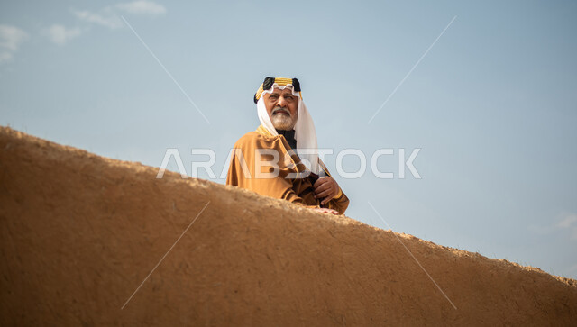 رجل مسن عربي سعودي خليجي يرتدي الزي السعودي الشعبي التقليدي, الزي التراثي للمناسبات الوطنيه, الوقوف في إحدى المباني الطينيه الشعبيه, بيوت تراثيه قديمه, شعبيات, يوم التأسيس السعودي, الإحتفالات الوطنيه في المنطقه الوسطى, مباني أثريه قديمه