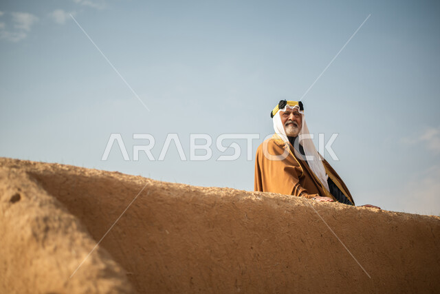 رجل مسن عربي سعودي خليجي يرتدي الزي السعودي الشعبي التقليدي, الزي التراثي للمناسبات الوطنيه, الوقوف في إحدى المباني الطينيه الشعبيه, بيوت تراثيه قديمه, شعبيات, يوم التأسيس السعودي, الإحتفالات الوطنيه في المنطقه الوسطى, مباني أثريه قديمه