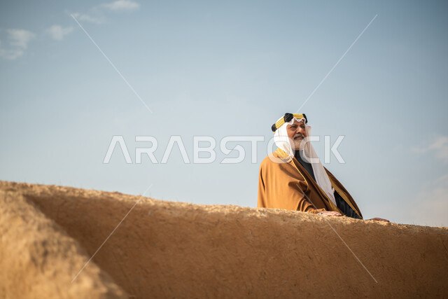رجل مسن عربي سعودي خليجي يرتدي الزي السعودي الشعبي التقليدي, الزي التراثي للمناسبات الوطنيه, الوقوف في إحدى المباني الطينيه الشعبيه, بيوت تراثيه قديمه, شعبيات, يوم التأسيس السعودي, الإحتفالات الوطنيه في المنطقه الوسطى, مباني أثريه قديمه