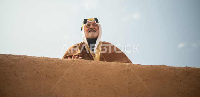 رجل مسن عربي سعودي خليجي يرتدي الزي السعودي الشعبي التقليدي, الزي التراثي للمناسبات الوطنيه, الوقوف في إحدى المباني الطينيه الشعبيه, بيوت تراثيه قديمه, شعبيات, يوم التأسيس السعودي, الإحتفالات الوطنيه في المنطقه الوسطى, مباني أثريه قديمه