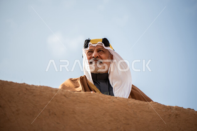 رجل مسن عربي سعودي خليجي يرتدي الزي السعودي الشعبي التقليدي, الزي التراثي للمناسبات الوطنيه, الوقوف في إحدى المباني الطينيه الشعبيه, بيوت تراثيه قديمه, شعبيات, يوم التأسيس السعودي, الإحتفالات الوطنيه في المنطقه الوسطى, مباني أثريه قديمه
