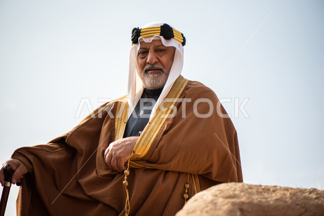 رجل مسن عربي سعودي خليجي يرتدي الزي السعودي الشعبي التقليدي, الزي التراثي للمناسبات الوطنيه, الوقوف في إحدى المباني الطينيه الشعبيه, بيوت تراثيه قديمه, شعبيات, يوم التأسيس السعودي, الإحتفالات الوطنيه في المنطقه الوسطى, مباني أثريه قديمه