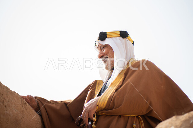 رجل مسن عربي سعودي خليجي يرتدي الزي السعودي الشعبي التقليدي, الزي التراثي للمناسبات الوطنيه, الوقوف في إحدى المباني الطينيه الشعبيه, بيوت تراثيه قديمه, شعبيات, يوم التأسيس السعودي, الإحتفالات الوطنيه في المنطقه الوسطى, مباني أثريه قديمه