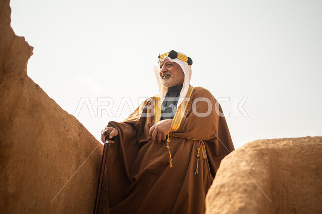 رجل مسن عربي سعودي خليجي يرتدي الزي السعودي الشعبي التقليدي, الزي التراثي للمناسبات الوطنيه, الوقوف في إحدى المباني الطينيه الشعبيه, بيوت تراثيه قديمه, شعبيات, يوم التأسيس السعودي, الإحتفالات الوطنيه في المنطقه الوسطى, مباني أثريه قديمه