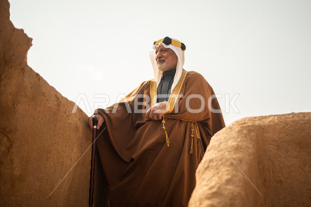 رجل مسن عربي سعودي خليجي يرتدي الزي السعودي الشعبي التقليدي, الزي التراثي للمناسبات الوطنيه, الوقوف في إحدى المباني الطينيه الشعبيه, بيوت تراثيه قديمه, شعبيات, يوم التأسيس السعودي, الإحتفالات الوطنيه في المنطقه الوسطى, مباني أثريه قديمه