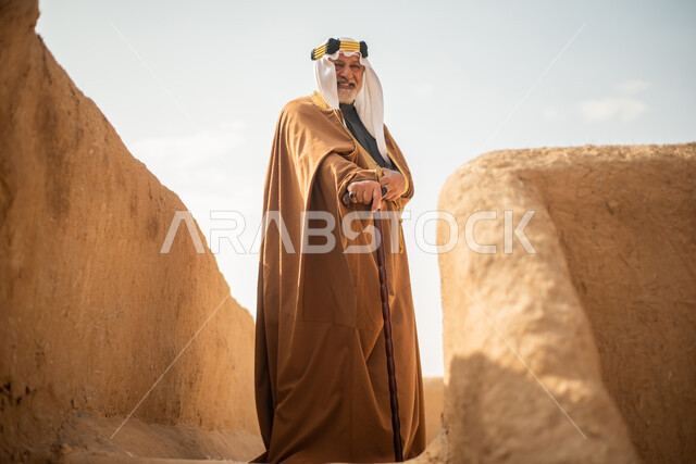 رجل مسن عربي سعودي خليجي يرتدي الزي السعودي الشعبي التقليدي, الزي التراثي للمناسبات الوطنيه, الوقوف في إحدى المباني الطينيه الشعبيه, بيوت تراثيه قديمه, شعبيات, يوم التأسيس السعودي, الإحتفالات الوطنيه في المنطقه الوسطى, مباني أثريه قديمه