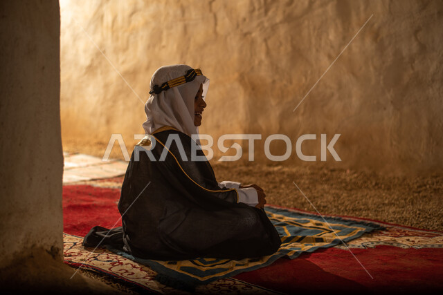 صوره من الخلف لولد عربي سعودي خليجي يرتدي الزي السعودي الشعبي التقليدي, الصلاه في إحدى المباني الطينيه الشعبيه, بيوت تراثيه قديمه, شعبيات