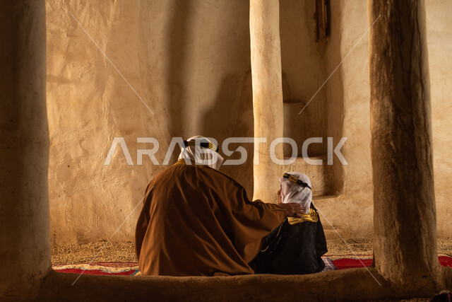 صوره من الخلف لرجل مسن وولد عربيان سعوديان خليجيان يرتديان الزي السعودي الشعبي التقليدي, الصلاه في إحدى المباني الطينيه الشعبيه, بيوت تراثيه قديمه, شعبيات
