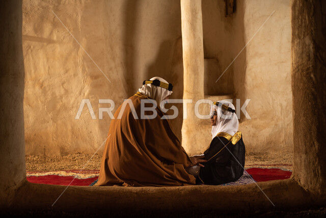 صوره من الخلف لرجل مسن وولد عربيان سعوديان خليجيان يرتديان الزي السعودي الشعبي التقليدي, الصلاه في إحدى المباني الطينيه الشعبيه, بيوت تراثيه قديمه, شعبيات