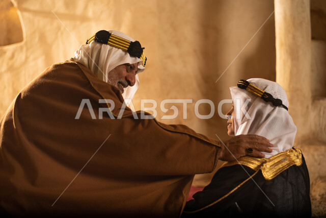 صوره من الخلف لرجل مسن وولد عربيان سعوديان خليجيان يرتديان الزي السعودي الشعبي التقليدي, الصلاه في إحدى المباني الطينيه الشعبيه, بيوت تراثيه قديمه, شعبيات