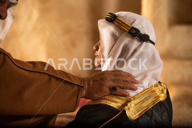 صوره من الخلف لرجل مسن وولد عربيان سعوديان خليجيان يرتديان الزي السعودي الشعبي التقليدي, الصلاه في إحدى المباني الطينيه الشعبيه, بيوت تراثيه قديمه, شعبيات