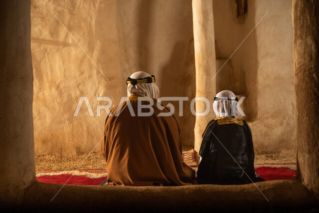 صوره من الخلف لرجل مسن وولد عربيان سعوديان خليجيان يرتديان الزي السعودي الشعبي التقليدي, الصلاه في إحدى المباني الطينيه الشعبيه, بيوت تراثيه قديمه, شعبيات