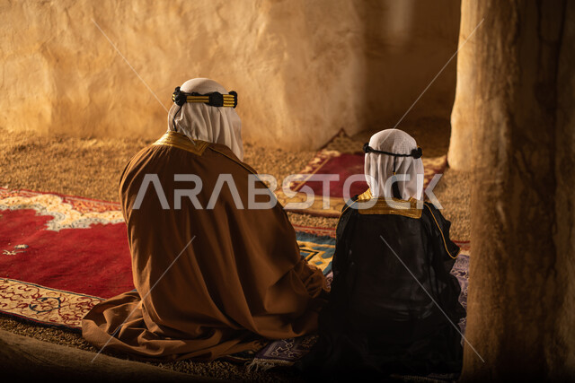 صوره من الخلف لرجل مسن وولد عربيان سعوديان خليجيان يرتديان الزي السعودي الشعبي التقليدي, الصلاه في إحدى المباني الطينيه الشعبيه, بيوت تراثيه قديمه, شعبيات