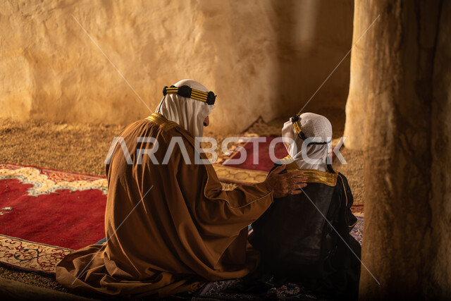 صوره من الخلف لرجل مسن وولد عربيان سعوديان خليجيان يرتديان الزي السعودي الشعبي التقليدي, الصلاه في إحدى المباني الطينيه الشعبيه, بيوت تراثيه قديمه, شعبيات