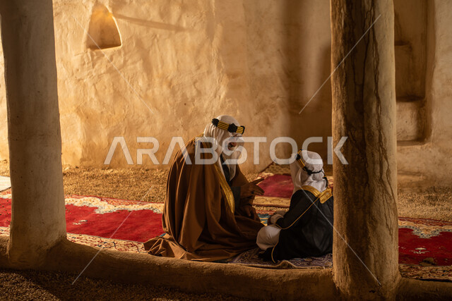 صوره من الخلف لرجل مسن وولد عربيان سعوديان خليجيان يرتديان الزي السعودي الشعبي التقليدي, الصلاه في إحدى المباني الطينيه الشعبيه, بيوت تراثيه قديمه, شعبيات