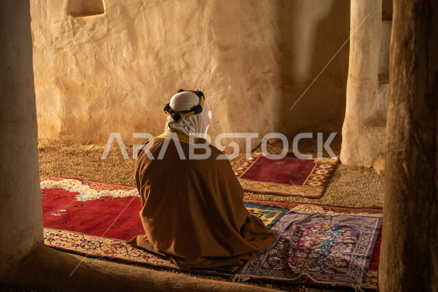 صوره من الخلف لرجل مسن عربي سعودي خليجي يرتدي الزي السعودي الشعبي التقليدي, الصلاه في إحدى المباني الطينيه الشعبيه, بيوت تراثيه قديمه, شعبيات
