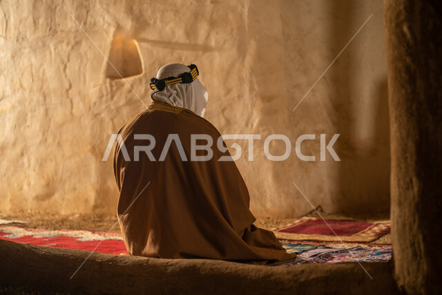 صوره من الخلف لرجل مسن عربي سعودي خليجي يرتدي الزي السعودي الشعبي التقليدي, الصلاه في إحدى المباني الطينيه الشعبيه, بيوت تراثيه قديمه, شعبيات