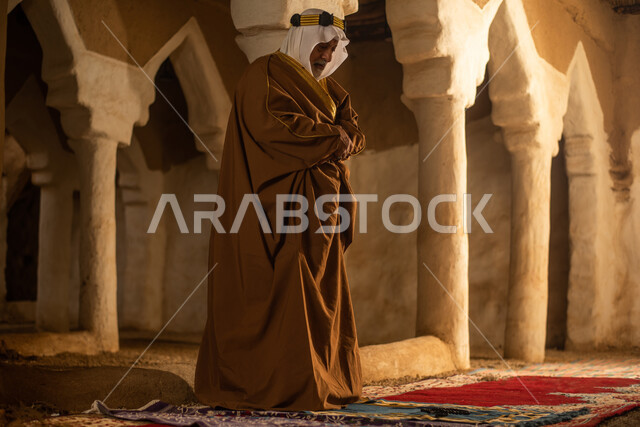 رجل مسن عربي سعودي خليجي يرتدي الزي السعودي الشعبي التقليدي, الصلاه في إحدى المباني الطينيه الشعبيه, بيوت تراثيه قديمه, شعبيات