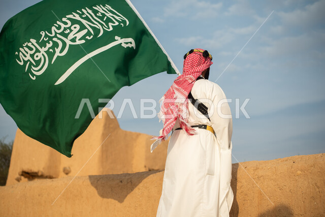 تصوير من الخلف لرجل عربي سعودي خليجي يرتدي الزي السعودي الشعبي التقليدي, الزي التراثي للمناسبات الوطنيه, الوقوف في إحدى المباني الطينيه الشعبيه, رؤيه مستقبليه، المستقبل السعودي، إيماءات النظر من بعيد, بيوت تراثيه قديمه, يوم التأسيس السعودي, مباني أثريه
