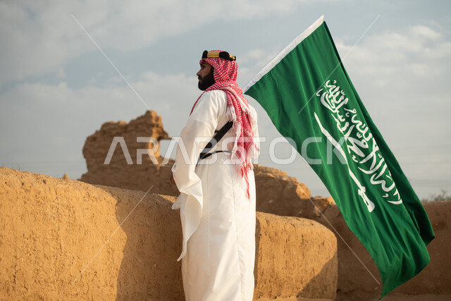 تصوير من الخلف لرجل عربي سعودي خليجي يرتدي الزي السعودي الشعبي التقليدي, الزي التراثي للمناسبات الوطنيه, الوقوف في إحدى المباني الطينيه الشعبيه, رؤيه مستقبليه، المستقبل السعودي، إيماءات النظر من بعيد, بيوت تراثيه قديمه, يوم التأسيس السعودي, مباني أثريه