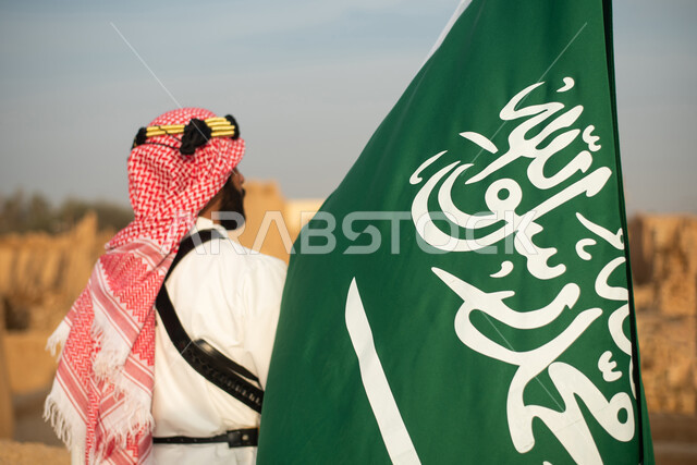 تصوير من الخلف لرجل عربي سعودي خليجي يرتدي الزي السعودي الشعبي التقليدي, الزي التراثي للمناسبات الوطنيه, الوقوف في إحدى المباني الطينيه الشعبيه, رؤيه مستقبليه، المستقبل السعودي، إيماءات النظر من بعيد, بيوت تراثيه قديمه, يوم التأسيس السعودي, مباني أثريه
