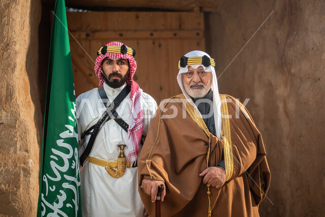 Two Saudi Arabian Gulf men dressed in the traditional Saudi folk costume, the traditional costume for national occasions, standing in one of the popular mud buildings, old heritage houses, folklore, Saudi founding day, national celebrations in the central