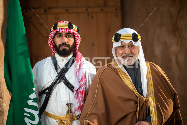 Two Saudi Arabian Gulf men dressed in the traditional Saudi folk costume, the traditional costume for national occasions, standing in one of the popular mud buildings, old heritage houses, folklore, Saudi founding day, national celebrations in the central