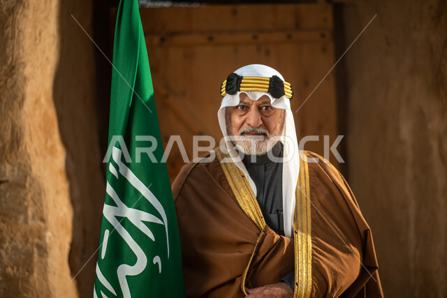 رجل مسن عربي سعودي خليجي يرتدي الزي السعودي الشعبي التقليدي, الزي التراثي للمناسبات الوطنيه, الوقوف في إحدى المباني الطينيه الشعبيه, بيوت تراثيه قديمه, شعبيات, يوم التأسيس السعودي, الإحتفالات الوطنيه في المنطقه الوسطى, مباني أثريه قديمه