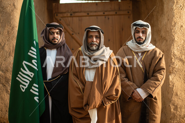 A group of Saudi Arab men, the flag of the Kingdom of Saudi Arabia, the Saudi founding day, the founding of the Kingdom in 1727 AD, Saudi folk costumes, old Saudi Arabia, old traditional mud houses, Saudi traditional costume for national occasions