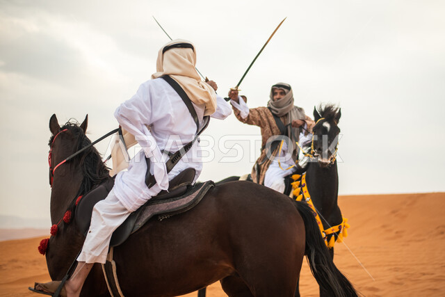 فارسان عربيان خليجيان سعوديان يمتطيان الخيل في الصحراء السعودية ويتبارزان، مبارزة السيوف، ارتداء زي تراثي شعبي، احياء ذكرى التاسيس السعودي، الدولة السعودية الاولى 1727م، يوم التاسيس السعودي، عيد وطني سعودي
