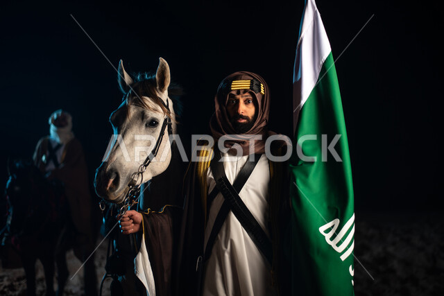 A Saudi Arab Gulf horseman wearing a traditional costume and standing with horses in the desert at night, raising the flag of Saudi Arabia, the purebred Arabian horse, commemorating the Saudi founding, the first Saudi state 1727 AD, the day of the Saudi f