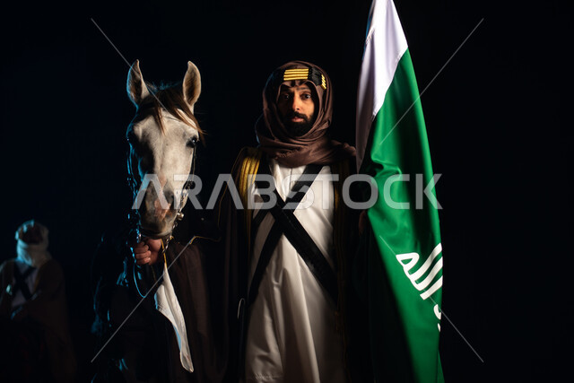 A Saudi Arab Gulf horseman wearing a traditional costume and standing with horses in the desert at night, raising the flag of Saudi Arabia, the purebred Arabian horse, commemorating the Saudi founding, the first Saudi state 1727 AD, the day of the Saudi f