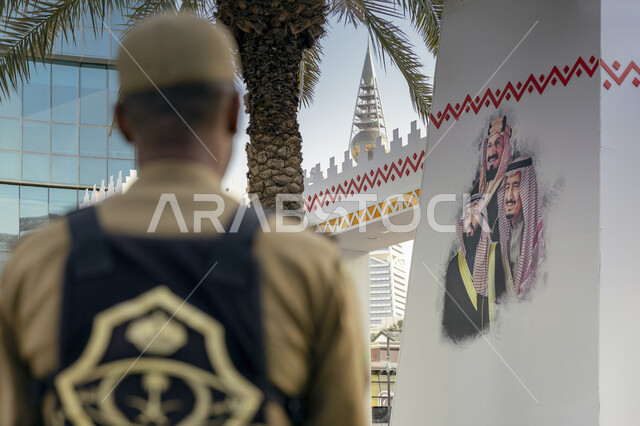 صورة من الخلف لرجل امن عربي خليجي سعودي، رسمة جدارية لمؤسس المملكة والملك سلمان وولي العهد، برج الفيصلية في الرياض، احياء ذكرى اليوم الوطني السعودي، يوم التاسيس السعودي