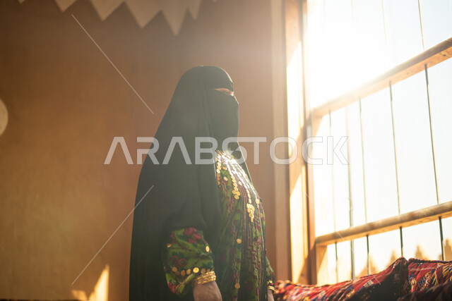 سيدة عربية خليجية سعودية ترتدي النقاب وتقف امام النافذة، ارتداء زي تراثي للمناسبات الوطنية والاحتفالية، البرقع السعودي، زي سعودي شعبي، النظر من خلال نافذة المنزل، البيوت الاثرية القديمة