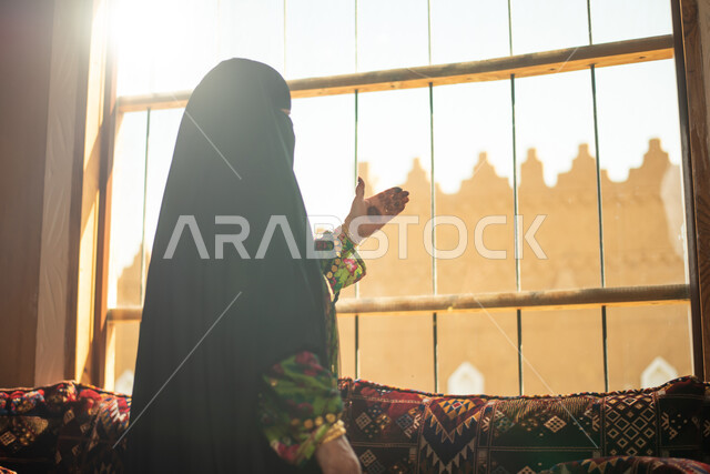 سيدة عربية خليجية سعودية ترتدي النقاب وتقف امام النافذة، ارتداء زي تراثي للمناسبات الوطنية والاحتفالية، البرقع السعودي، زي سعودي شعبي، الرسم بالحناء على كف اليد، النظر من خلال نافذة المنزل، البيوت الاثرية القديمة