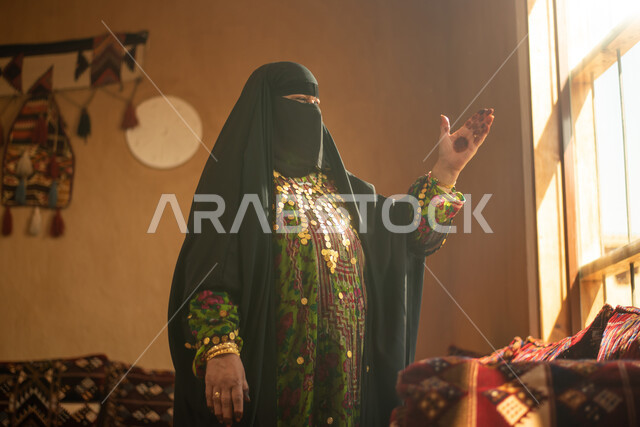 سيدة عربية خليجية سعودية ترتدي النقاب وتقف امام النافذة، ارتداء زي تراثي للمناسبات الوطنية والاحتفالية، البرقع السعودي، زي سعودي شعبي، الرسم بالحناء على كف اليد، النظر من خلال نافذة المنزل، البيوت الاثرية القديمة