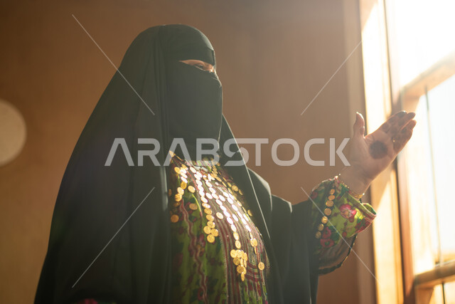 سيدة عربية خليجية سعودية ترتدي النقاب وتقف امام النافذة، ارتداء زي تراثي للمناسبات الوطنية والاحتفالية، البرقع السعودي، زي سعودي شعبي، الرسم بالحناء على كف اليد، النظر من خلال نافذة المنزل، البيوت الاثرية القديمة