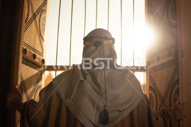 صورة من الخلف لرجل عربي خليجي سعودي يرتدي زي تراثي للمناسبات الوطنية والاحتفالية ويقف امام نافذة المنزل، زي سعودي شعبي، النظر من خلال النافذة، بيوت قديمة، نافذة خشبية