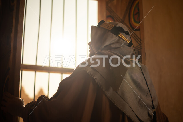صورة من الخلف لرجل عربي خليجي سعودي يرتدي زي تراثي للمناسبات الوطنية والاحتفالية ويقف امام نافذة المنزل، زي سعودي شعبي، النظر من خلال النافذة، بيوت قديمة، نافذة خشبية