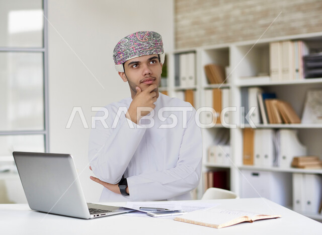 رجل عربي عماني خليجي باللباس التقليدي , يقوم بإشارات وجه تدل على التفكير والتركيز, إستخدام جهاز الحاسوب المحمول، تصفح الإنترنت، جلسة عمل، العمل عن بعد ، التسويق الإلكتروني، إجراء مكالمة فيديو .